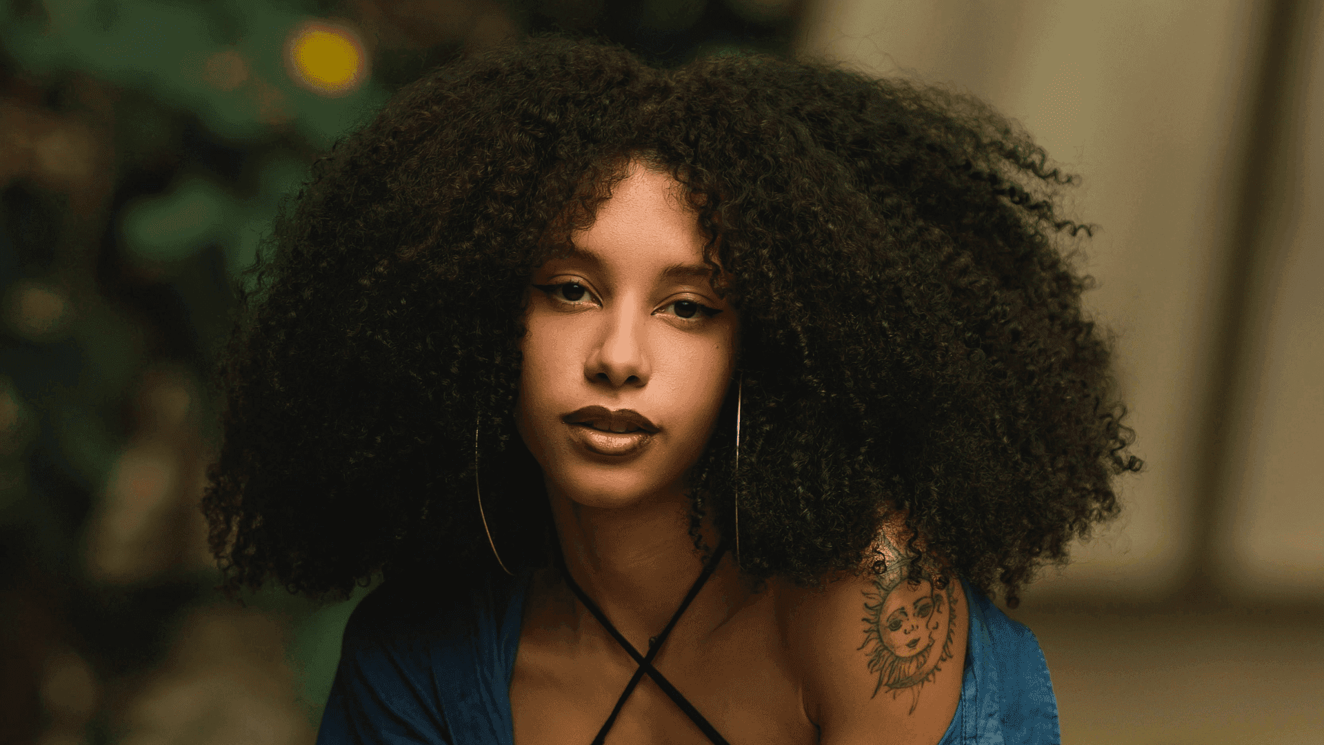 Young woman with curly hair and a sun tattoo, wearing hoop earrings.