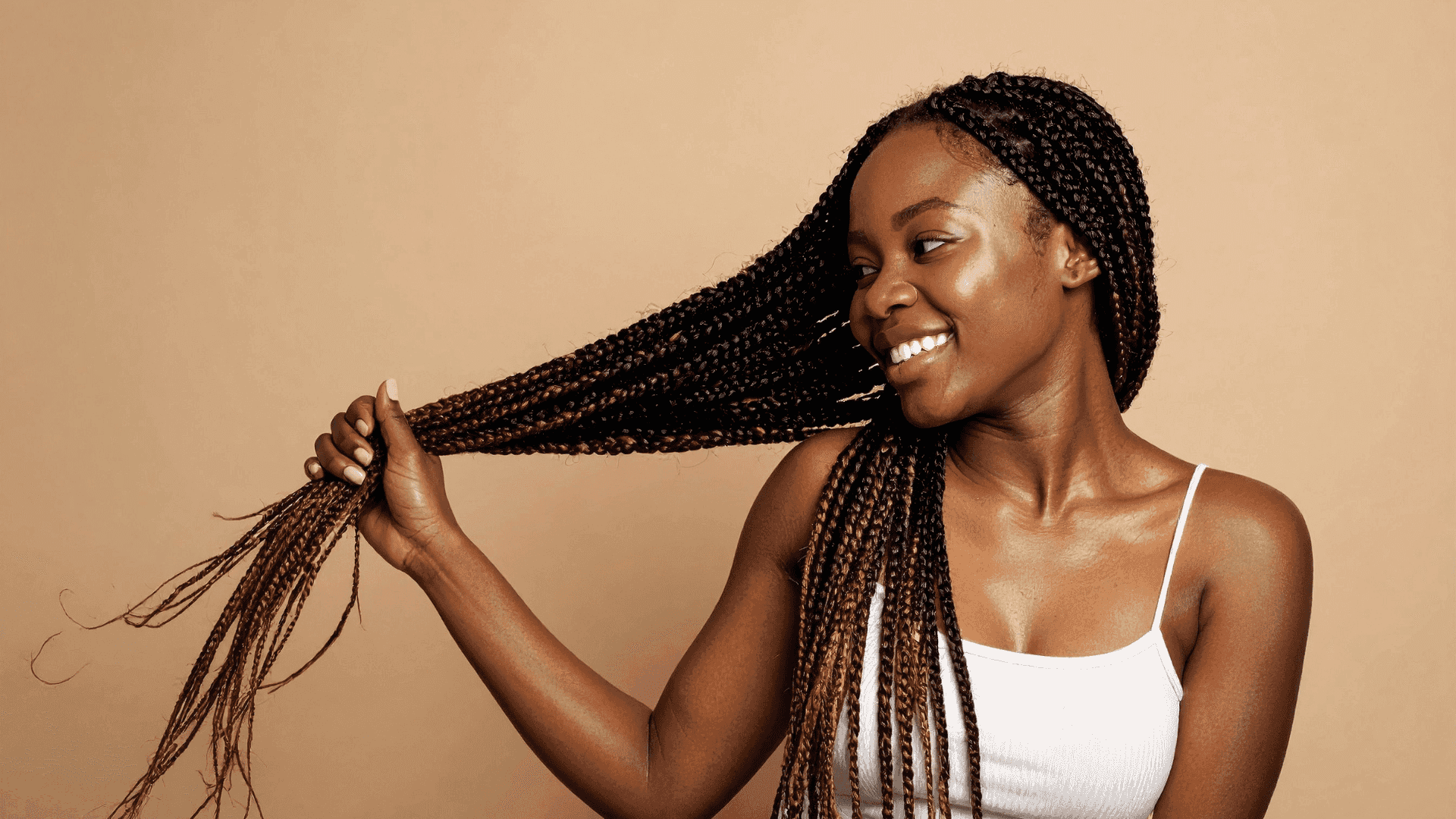 A woman in a white tank top smiling, holding her long braided hair against a beige backdrop.